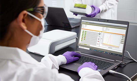 A lab technician in a white coat and purple gloves opens a Neogen molecular detection system for pathogen testing on a laboratory bench.